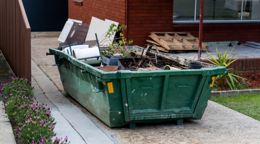 a green roll-off dumpster in a driveway, full of household items and yard waste